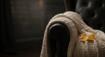 A cozy cream-colored knitted blanket with a golden maple leaf draped over a dark brown leather armchair, creating a warm and inviting autumn scene indoors.