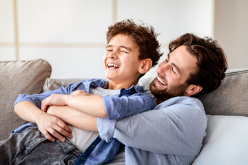 Family weekend. Loving dad tickling his laughing son, spending leisure time at home together, sitting on sofa and smiling. Fatherhood concept, relationship with daddy