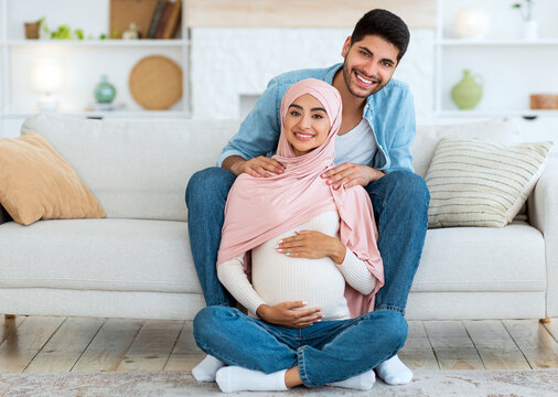 Massage during pregnancy concept. Young arab man massaging his wife's shoulders, resting together at home. Positive muslim gyt taking care of his expectant girlfriend