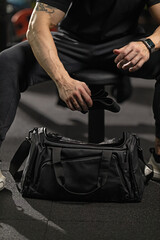 Muscular man sits beside a bench press barbell in a gym reaching into his bag to pull out wrist wraps. He is focused on getting ready for an intense lifting session.