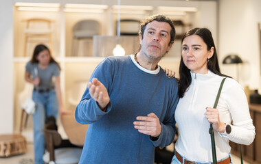 Obraz premium Husband and wife are discussing the purchase of new furniture for the house, they are considering furniture made of natural wood. European couple choose chairs and a sofa for an apartment after