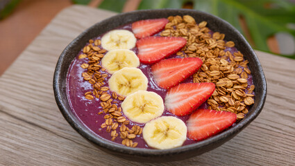 A visually appealing healthy smoothie bowl sits on a rustic wooden table. The bowl is filled with a thick, purple acai base and garnished with fresh slices of banana and strawberry.