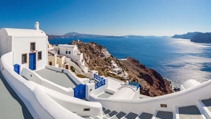 Obraz premium A panoramic view of the iconic white houses and blue domes of Santorini, Greece. The buildings are perched on a cliffside, overlooking the deep blue caldera and the Aegean Sea, with the sky clear.