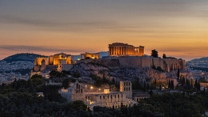 A stunning view of the Acropolis and the Parthenon in Athens, Greece, during sunset or dusk. The ancient ruins are beautifully illuminated with golden light, standing proudly on a rocky hill.