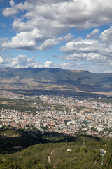 scenic view of a city nestled in a valley, surrounded by rolling green hills and distant mountains under a cloudy blue sky