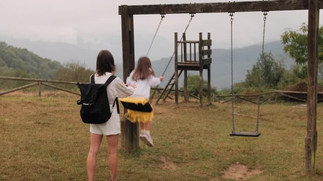 Rear view of a mother pushing her young daughter on a wooden swing at a rural hillside playground on an overcast day.