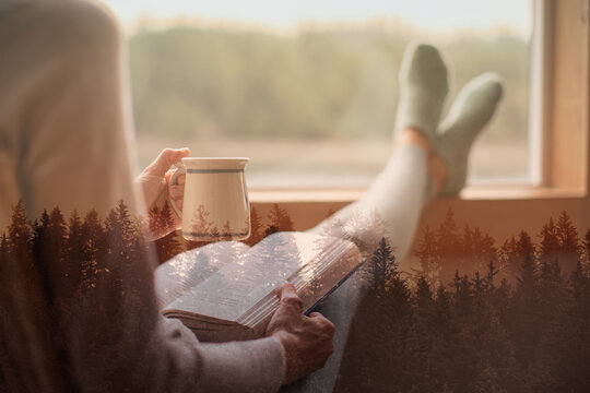 Tranquil moment of relaxation featuring a cozy woman reading a book, sipping a hot beverage, and enjoying the serene view of nature with a double exposure effect of forest scenery.
