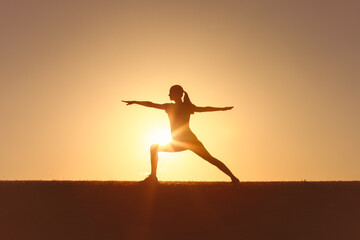 Fototapeta premium Woman in a yoga stretching exercise pose during sunset, symbolizing balance and harmony. Silhouette against the glowing orange sky conveys inspiration and tranquility in outdoor fitness activities 