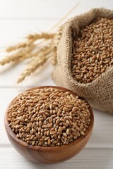 Wheat grains in bowl, burlap bag and spikelets on white wooden table, closeup