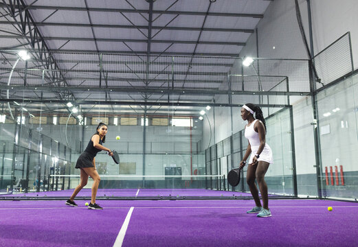 Diverse female opponents swinging padel rackets, returning yellow ball over net on purple court