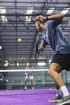 Male opponents hitting yellow padel ball using rackets at net on purple court indoors
