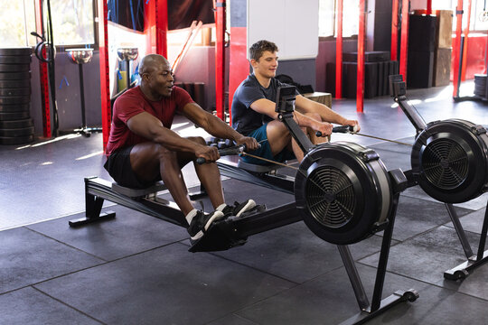 Diverse male workout partners rowing on machines in gym wearing athletic t-shirts, shorts