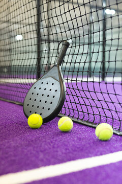 Black padel racket leaning against black net on purple court, three balls resting nearby