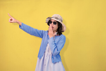 Elegant young woman wearing a stylish hat and sunglasses exhibiting surprise on a yellow backdrop