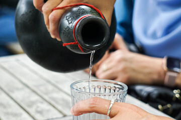 Hands pouring clear liquid from black ceramic bottle into glass on rustic table, capturing a serene outdoor moment with cultural elegance.