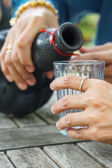 Hands pouring clear liquid from black ceramic bottle into glass on rustic table, capturing a serene outdoor moment with cultural elegance.