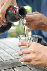 Hands pouring clear liquid from black ceramic bottle into glass on rustic table, capturing a serene outdoor moment with cultural elegance.