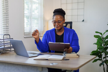 Portrait of a cheerful excited happy businesswoman sitting at desk in modern office. Smiling african american executive using laptop while working from office with copy space.
