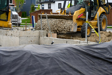 Front loader heavy construction equipment with bucket full of gravel dumping it into a new house foundation, new housing development under construction
