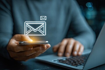 Person holding phone with email icon overlay, laptop in the background