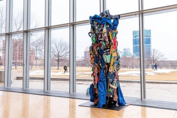 Tall, free-standing sculpture composed of layered, colorful fabric scraps, displayed indoors near large windows overlooking a park