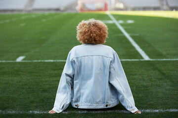 Young person with curly hair wearing a denim jacket sits on the sidelines of a football field looking out