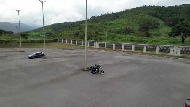 Drone shot, zoom out a lone biker and their motorcycle alongside rest of the road with lush vegetation Background