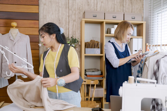 Young asian fashion designer holding garment and hanger while preparing clothing display in home studio for handmade apparel business with organized textile workspace and dress form in background