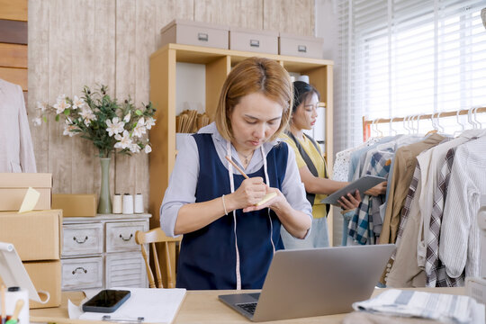 Two asian female designers discussing packaging while managing small fashion business from home studio workspace with fabric, sewing tools, parcels, and modern branding environment