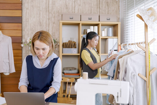 Asian adult female fashion designer working laptop beside sewing machine while another designer selecting clothes on rack in bright modern home studio small business workspace