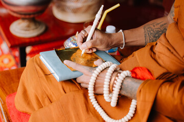 An elderly person's hands hold a white thread, likely part of a Buddhist practice, with an orange robe in the background.