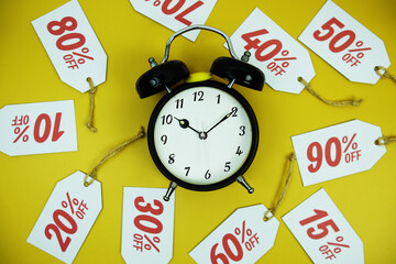 Clock surrounded by various discount tags with percentages like 10%, 20%, 30%, up to 90% off, set against a bright yellow background