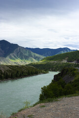 Turquoise waters of the Katun River wind through lush green valleys and rugged cliffs of Altai Mountains beneath cloudy summer sky. Scenic view of Katun River in Altay