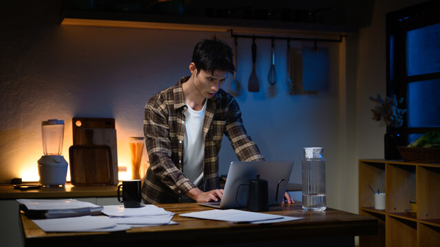 Focused man working late at night in kitchen, typing on laptop with documents on table