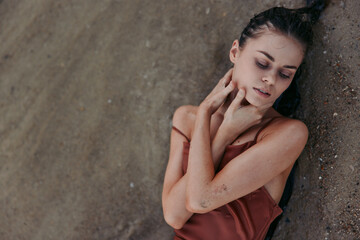 A thoughtful woman rests against a textured wall, eyes closed, chin tilted, and fingers brushing her neck. Soft lighting highlights her serene, introspective mood.
