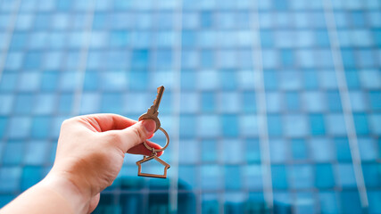 Close-up of hand holding house key with keychain in front of modern glass building. Concept of real estate, new home ownership, apartment investment, property rental, and urban lifestyle.