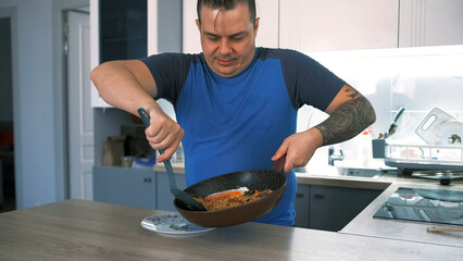 A fat guy has cooked himself a meal and is putting hot food from a frying pan on his plate. A male...