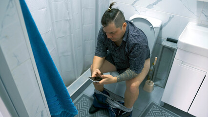 A man with his underpants and trousers down is sitting on the toilet and looking at his mobile phone. The guy is sitting in the toilet