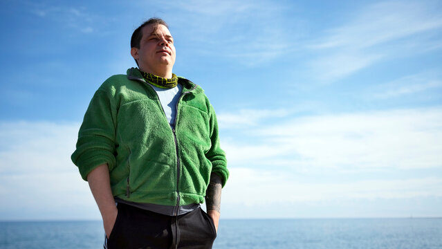 A happy, confident, obese guy in a green sweatshirt and black pants stands against a background of blue sky and sea. The man enjoys the view