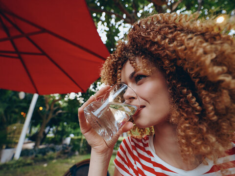 A young woman with curly hair drinks water from a glass outdoors, capturing a refreshing moment in warm light, wearing a casual striped shirt, and posing in a natural candid portrait.