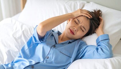 A young woman in blue pajamas lies in bed, clutching her head, eyes closed, looking distressed, suggesting a headache or illness