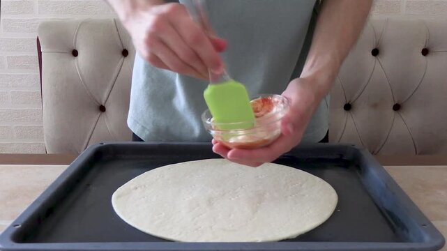 Greasing the pizza dough with the sauce using a silicone spatula. Pizza dough on a baking sheet