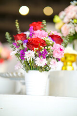 colorful flower arrangement in a white vase. The bouquet includes red roses, pink and purple flowers, and green leaves, placed on a white surface with a blurred background.