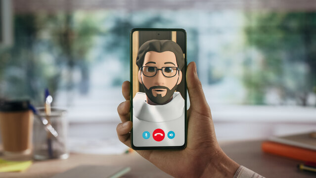 Close-up of Hand Holding Smartphone During Video Call With 3d Avatar. Animated Character With Glasses and Beard on Screen. Blurred Home Office Background With Desk Items and Stationery.