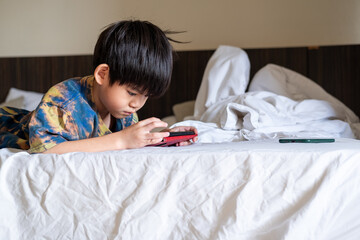 Asian boy lying on bed happily playing with mobile phone enjoying games and digital content