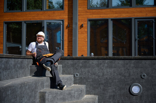 Construction worker relaxing near empty swimming pool. Builder in uniform has a break sitting in spa zone. Man resting at construction site. Tired building contractor. Summer day. Lazy engineer - Powered by Adobe