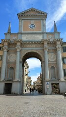 Obraz premium A monumental archway with a clock and classical columns, located in a cobblestone square in the historic town of Bra, Italy, under a blue sky.