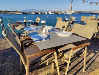 Serene waterfront café with yellow chairs and tables overlooking a calm sea, framed by boats and distant buildings under a clear sky. Metaphorically, it symbolizes a haven of rest, connection.