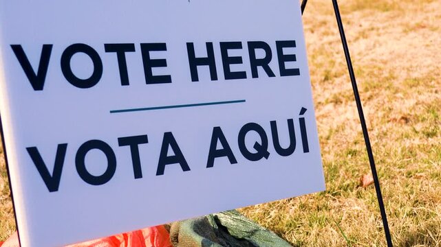 Vote Here Vota Aqu&iacute; sign in close-up with bold bilingual lettering, standing out clearly as cars drive past in the blurred background. Imagery for election day, civic duty, democracy and participation