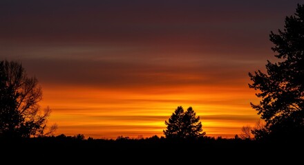 Naklejka premium A vibrant sunset paints the sky with fiery orange and deep purple hues. Silhouetted trees frame the dramatic, layered cloudscape